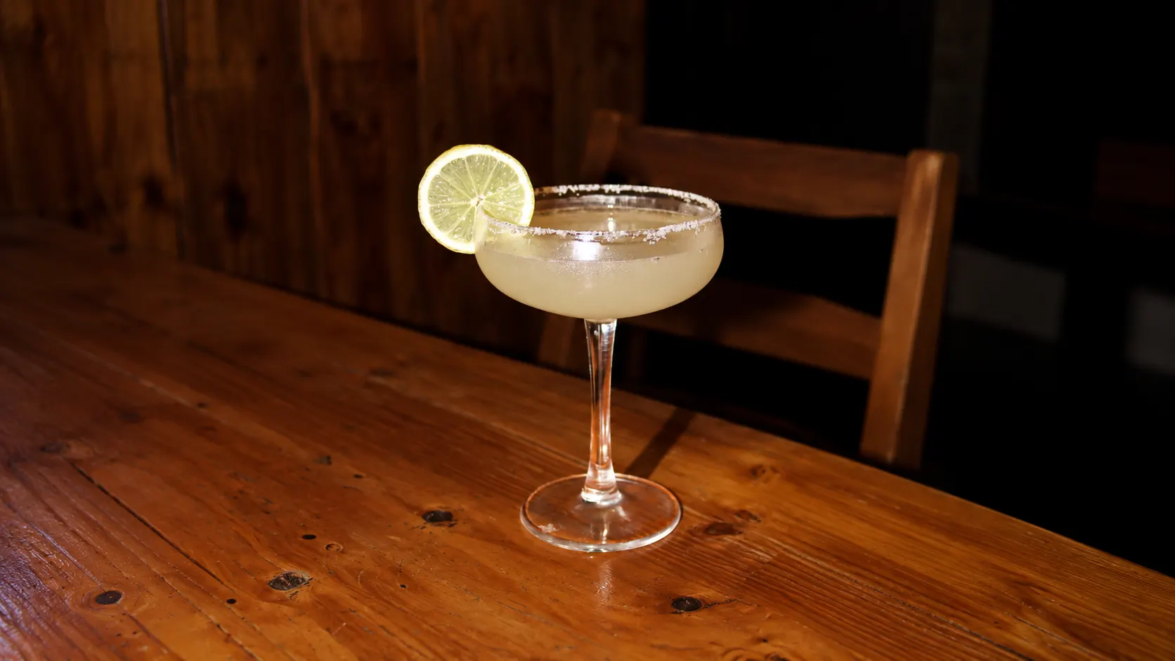 Uma margarita numa taça com sal no rebordo e uma rodela de lima, numa mesa de madeira.