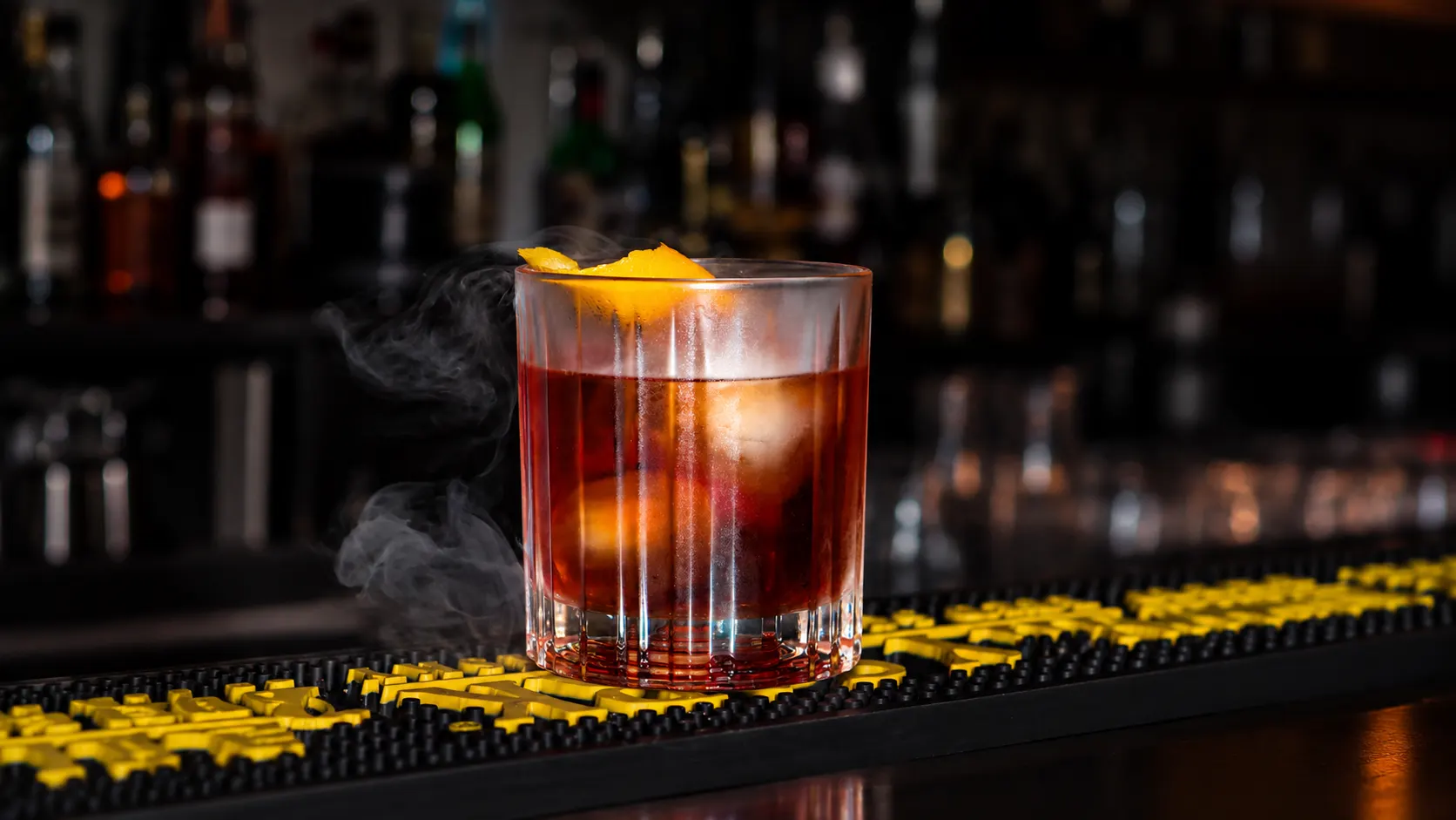 Um negroni com casca de laranja, envolto em fumo, em cima do balcão.