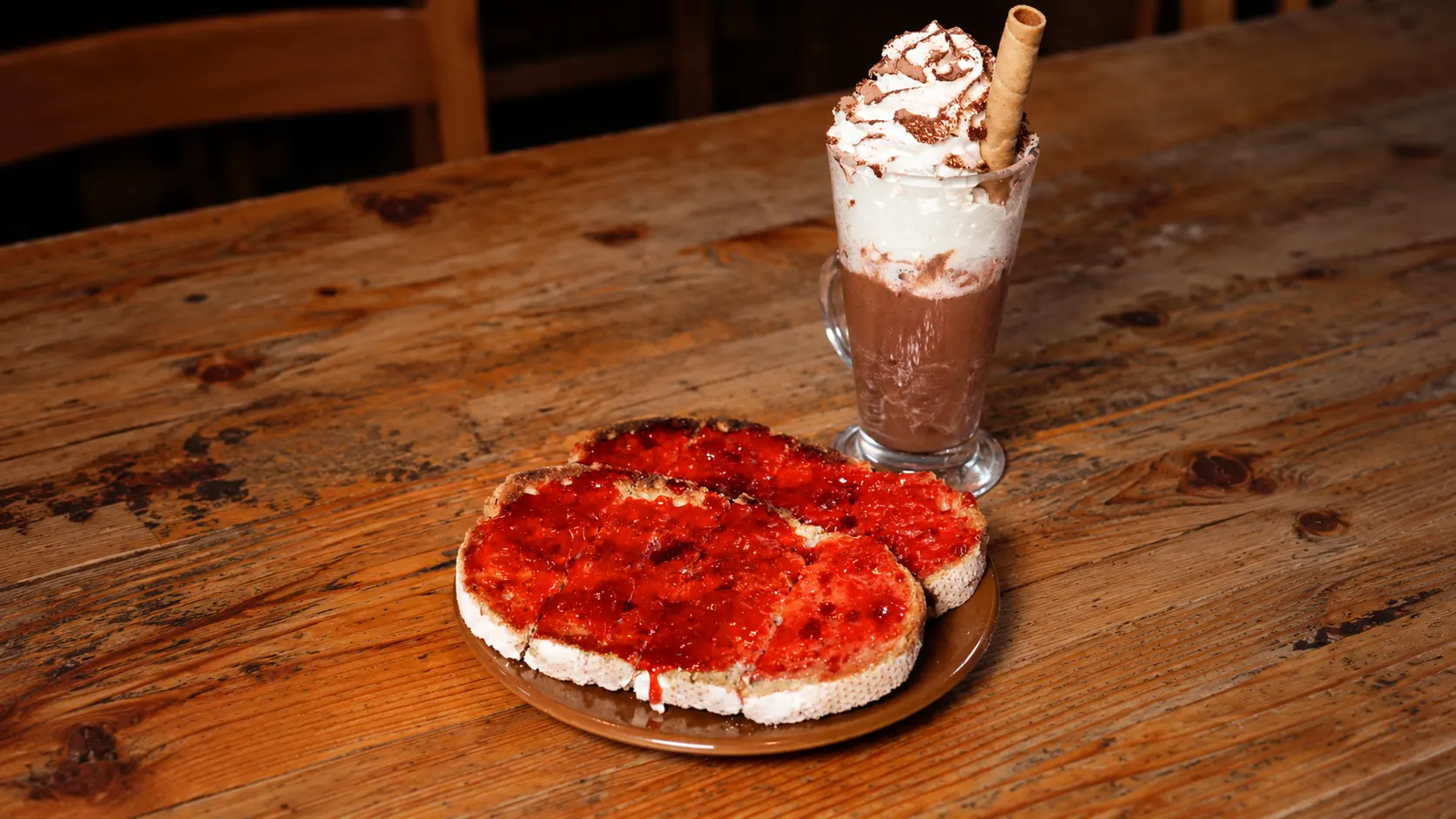 Um chocolate quente com chantilly e um barquilho, atrás de duas torradas com compota, numa mesa de madeira.